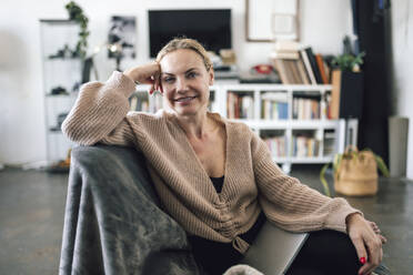 Smiling woman sitting with laptop on armchair at home - RFTF00225