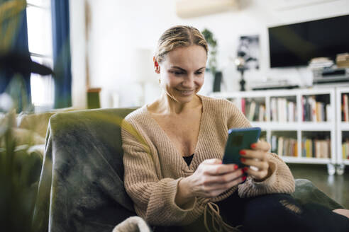 Smiling woman using mobile phone sitting on armchair at home - RFTF00226