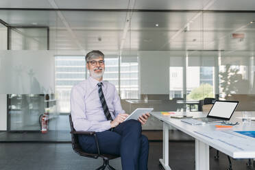 Smiling businessman with tablet PC sitting on chair in office - MEUF05762