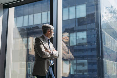 Businessman with arms crossed looking through window in office - MEUF05776