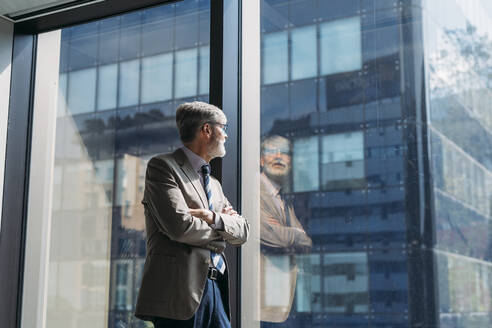 Geschäftsmann mit verschränkten Armen schaut durch ein Fenster im Büro - MEUF05776