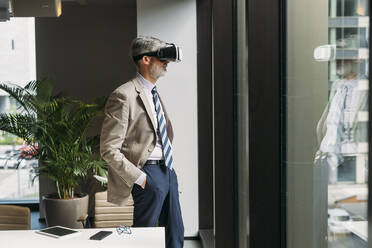Geschäftsmann, der einen Virtual-Reality-Simulator trägt und mit den Händen in den Taschen am Fenster im Büro steht - MEUF05787
