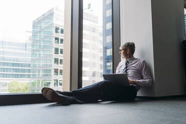 Geschäftsmann mit Laptop sitzt auf dem Boden am Fenster im Büro - MEUF05800