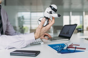 Hand of businessman holding toy robot sitting at desk at office - MEUF05813