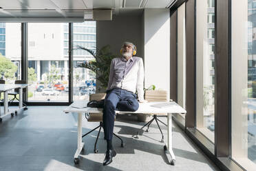 Businessman wearing wireless headphones sitting on desk in office - MEUF05818