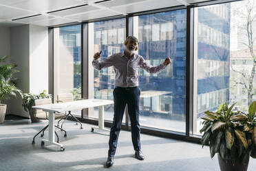 Businessman listening music through wireless headphones dancing in front of window at office - MEUF05820