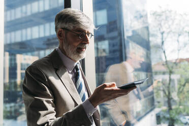 Happy businessman using smart phone standing by window in office - MEUF05824