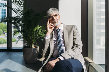 Mature businessman wearing eyeglasses sitting on chair by window at office - MEUF05827