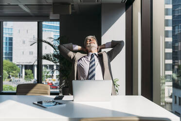 Businessman with hands behind head sitting at desk in office - MEUF05828