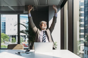 Happy businessman sitting with arms raised at desk in office - MEUF05829