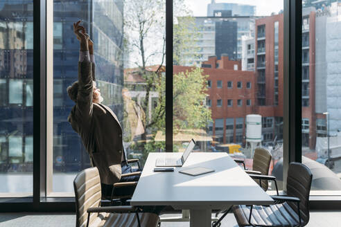 Businessman stretching arms sitting at desk by window in office - MEUF05830