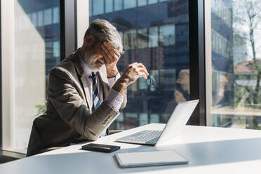 Tired businessman sitting with hand covering eye at desk in office - MEUF05831