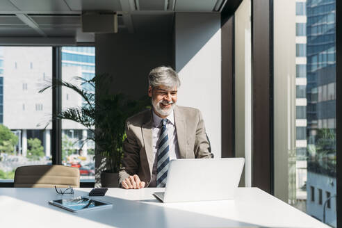 Glücklicher Geschäftsmann mit Laptop am Fenster sitzend am Schreibtisch im Büro - MEUF05833