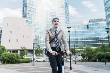 Businessman riding electric push scooter in front of modern office buildings - MEUF05839