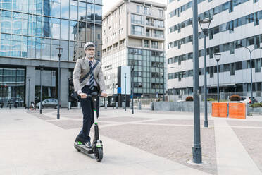 Mature businessman riding electric push scooter in front of modern office buildings - MEUF05845