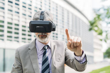 Älterer Geschäftsmann, der einen Virtual-Reality-Simulator trägt und vor einem Gebäude gestikuliert - MEUF05856
