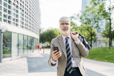 Lächelnder Geschäftsmann mit Smartphone vor einem Bürogebäude stehend - MEUF05864