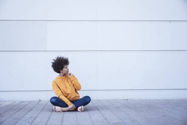 Young woman looking away sitting cross-legged in front of wall - UUF26201