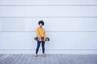 Young woman holding skateboard standing in front of wall - UUF26212