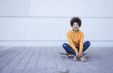 Smiling woman sitting cross-legged on skateboard in front of wall - UUF26213