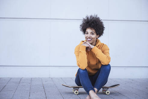 Lächelnde Afro-Frau sitzt auf einem Skateboard vor einer Wand - UUF26214