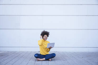 Woman on video call through tablet PC sitting cross-legged in front of wall - UUF26228