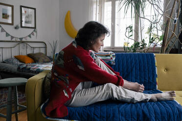 Side view of worried senior woman sitting on sofa examining foot at home - MASF29723