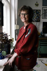 Smiling senior woman wearing eyeglasses standing near plants at home - MASF29724