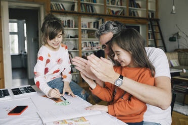 Glücklicher Vater gestikuliert, während er seiner Tochter bei den Hausaufgaben hilft, während das Mädchen am Tisch zeichnet - MASF29749
