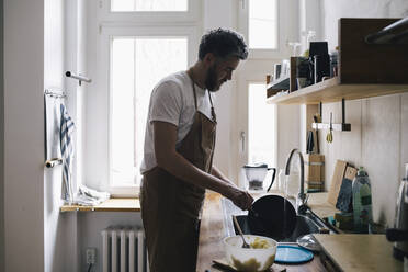 Side view of man wearing apron washing dishes in kitchen at home - MASF29761