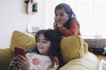 Mädchen mit Kopf in den Händen, die ihre Schwester beim Telefonieren beobachtet, während sie auf einem Sessel im Wohnzimmer sitzt - MASF29766