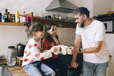 Lächelnder Vater, der mit seinen Zwillingstöchtern auf dem Küchentisch zu Hause Tassen anstößt - MASF29784