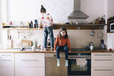 Mädchen in voller Länge, das neben seiner Schwester sitzt und im Regal sucht, während es auf der Küchentheke zu Hause steht - MASF29786