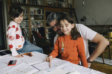 Smiling father looking at daughter reading book while girl sitting on table - MASF29793