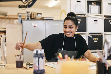 Lächelnde Studentin in Schürze malt am Tisch im Kunstunterricht - MASF29813
