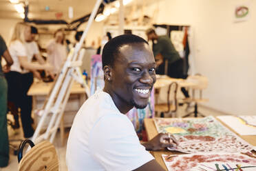 Porträt eines glücklichen männlichen Künstlers, der mit Gemälden an einem Tisch im Kunstunterricht sitzt - MASF29837
