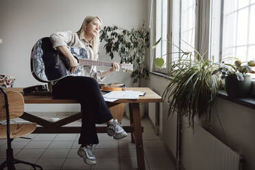 Reife Frau spielt Gitarre und sitzt auf einem Tisch im Klassenzimmer - MASF29847