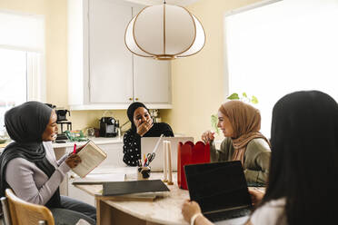 Fröhliche junge Frauen mit Hijabs sitzen am Tisch in der Küche - MASF29917