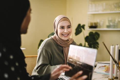 Lächelnde junge Frau im Hidschab sitzt bei einem Freiberufler mit Laptop zu Hause - MASF29922