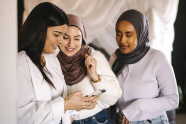 Lächelnde junge Frauen teilen ihr Smartphone mit Freunden zu Hause - MASF29967