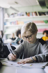 Schoolboy writing in book while sitting at desk in classroom - MASF30002