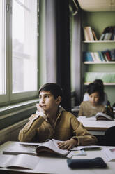 Thoughtful boy with hand on chin looking outside window while sitting in classroom - MASF30005