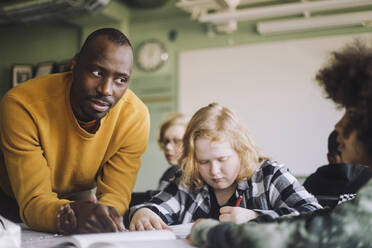 Ernster Lehrer, der einen Schüler ansieht, während er sich an den Schreibtisch im Klassenzimmer lehnt - MASF30059