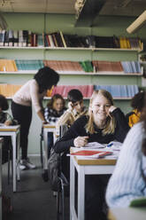 Smiling girl leaning on elbows while sitting at desk in classroom - MASF30077