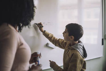 Seitenansicht eines Jungen, der mit einem Lehrer im Klassenzimmer eine mathematische Aufgabe auf einem weißen Brett löst - MASF30094