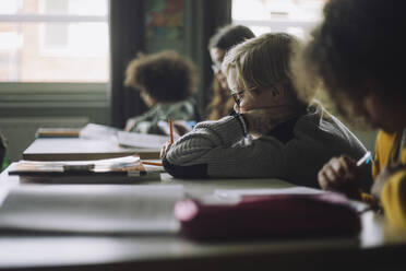 Seitenansicht eines Jungen beim Schreiben, der sich im Klassenzimmer auf den Ellbogen stützt - MASF30102