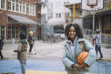 Portrait of smiling girl with basketball standing in sports court at school campus - MASF30119