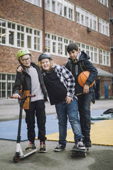 Boy holding push scooter with friends standing on skateboard in front of school building - MASF30124