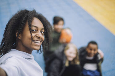 Glückliches Mädchen mit lockigem Haar nimmt Selfie mit Freunden im Hintergrund auf Sportplatz - MASF30130