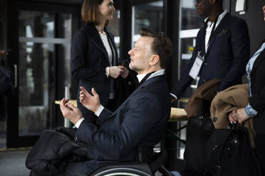 Side view of disabled businessman gesturing while sitting in wheelchair by delegates at seminar - MASF30177